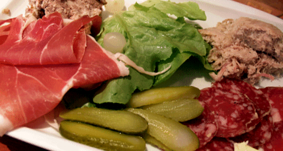 A plate of various cold cut meat, meat spreads, lettuce, and pickle slices.
