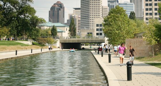 Indiana Central Canal with people walking along the sidewalk and a canal bridge with city buildings in the background.